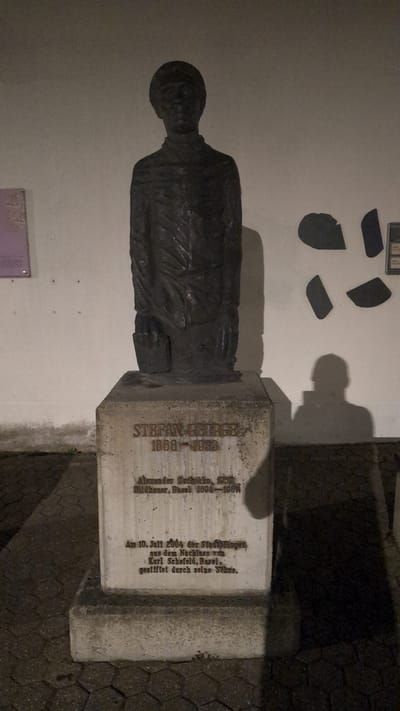 This is a medium shot, eye-level, outdoor photograph taken at night of a bronze bust of Stefan George (1868-1933) located in Bingen am Rhein, Germany. The bust is mounted on a large, light-colored concrete pedestal. The pedestal has an inscription that reads "STEFAN GEORGE 1868-1933" in large capital letters. Below this, there is smaller text that reads "Alexander Zschokke, 1857- Bildhauer, Basel 1894-1968N" and then below that "Am 10. Juli 2004 der Stadt Bingen aus dem Nachlass von Karl Schefold, Basel, gestiftet durch seine Söhne." This translates to "Alexander Zschokke, 1857- Sculptor, Basel 1894-1968N" and "On July 10, 2004, the City of Bingen from the estate of Karl Schefold, Basel, donated by his sons."
The bust depicts a man wearing a collared shirt and a jacket, with his left hand appearing to hold a book or document. The statue is dark and has a rough, textured finish. The background consists of a light-colored wall with some abstract shapes on it and shadows cast by the statue and an unseen person holding a phone. The ground in front of the pedestal is paved with hexagonal stones. The lighting suggests artificial illumination from above, casting strong shadows. The overall scene is stark and solemn.