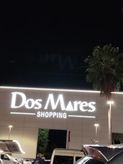 A nighttime shot showcases the "Dos Mares SHOPPING" sign illuminated against the building's facade. The sign is modern, with clean lines and bold lettering. A palm tree stands to the right of the building. Below the sign, a doorway is visible, with hints of greenery inside. Parked vehicles are in the foreground, and a few people can be seen near the entrance. The sky is dark. There seems to be some writing or a logo faded into the top part of the picture.
