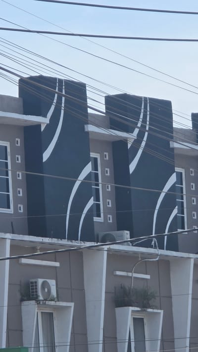 The image shows an upward angled view of two buildings in Meureudu, Indonesia. The building on the left is characterized by dark grey balconies adorned with curved white stripes, with visible electrical wires running vertically in front of it. Each level features windows with white frames, and small square architectural details above them. The building on the right has projecting white angled ledges, some of which contain air conditioning units and small potted plants. Both buildings have a neutral greyish-brown facade. The sky is pale blue, visible in the upper left corner. The overall scene captures a slice of the urban environment with a mix of architectural styles and utilities.