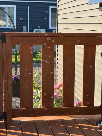 The image depicts an outdoor scene, likely a backyard or deck area, in Dieppe, Canada. In the foreground, a wooden deck and railing are visible, with sunlight casting shadows across the wooden planks. Numerous iridescent bubbles are scattered throughout the frame, some floating in the air and others resting on surfaces. Behind the railing, a patch of gravel and green foliage can be seen. Further back, the siding of a house is visible, featuring horizontal light-colored panels. To the left, the dark blue siding of another section of the house is present, with white-framed windows and a white downspout. An air conditioning unit is mounted on the wall of the light-colored house. The overall impression is one of a calm, sunny day with children's activities suggested by the presence of bubbles.