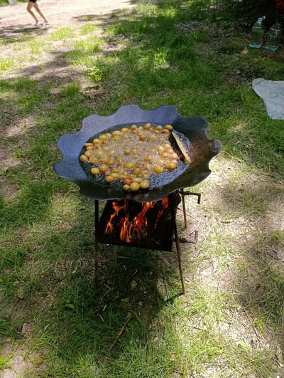 This is a high-angle, full shot of a large, black, scalloped pan filled with small, golden-brown potatoes being fried over an open fire. The pan rests on a simple metal grill, beneath which flames are visible, indicating an active fire. A wooden utensil, possibly a spatula or stirring stick, is partially submerged in the oil among the potatoes. The scene is set outdoors on a grassy area, with patches of dirt and fallen leaves visible. Sunlight casts shadows from trees onto the ground, suggesting a shaded location. In the upper left background, a person's legs are visible, walking on a dirt path. In the upper right background, two large plastic bottles, possibly filled with water, and a white cloth are placed on the grass. The overall impression is of an outdoor cooking event or picnic. The location is not specified as Capriana, Moldova, and there is no information in the image to support this claim.