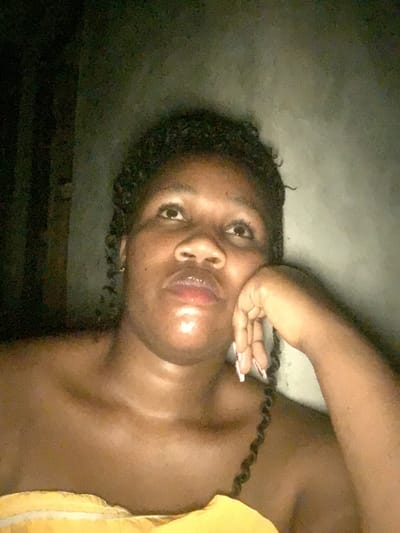 A dark-skinned woman poses with her hand on her cheek, showcasing long, painted fingernails. She has braided hair and shiny lips. Part of a yellow garment is visible in the lower right corner. The background is mostly dark and underexposed, creating a contrast with the subject's skin. The image appears to have been taken in low light or deliberately darkened.