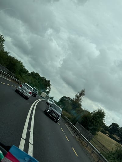 A traffic jam is depicted on a road in Scartnamore, Ireland. The overcast sky is filled with grey clouds. Several cars, including a silver Volkswagen hatchback, a black Volkswagen SUV, and a white van, are stopped on the asphalt road, which is marked with white and yellow lines. Trees and hedges line the side of the road, and in the distance, a field with a fence is visible. The perspective suggests the photo was taken from inside another vehicle.