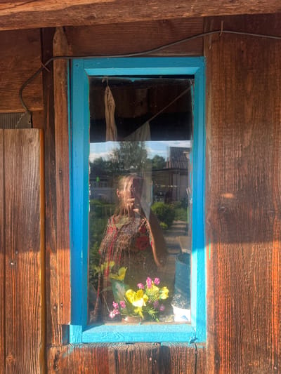 The media file shows a close-up, slightly angled view of a rustic wooden wall with a small, blue-framed window. The wall is constructed from weathered, horizontal wooden planks, with visible knots and nails. A black wire runs vertically along the left side of the wall, secured with a small metal staple.

Through the window, reflections are prominent, obscuring a clear view of the interior. A person's face, appearing to be a woman with blonde hair, is reflected in the glass. She is wearing a patterned top with red and green elements. Also reflected is a colorful arrangement of artificial flowers, including yellow and pink blossoms, placed inside.

The scene outside the window is partially visible through the reflections and the glass itself. It appears to be a sunny day, with bright daylight casting shadows on the wooden wall, particularly a diagonal shadow across the top right section. Trees and structures can be vaguely discerned in the background, suggesting an outdoor setting, possibly a rural or semi-rural location like Пигозино, Russia. The overall impression is one of a simple, weathered structure bathed in warm sunlight. There is no visible text in the image.