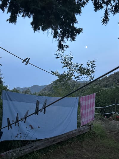 The image depicts an outdoor scene with laundry hanging on a clothesline, set against a backdrop of rolling hills. A light blue checkered sheet is prominently featured on the left, secured by several wooden clothespins. To its right, a pink and white checkered towel hangs, also clipped to the line. The clothesline itself is strung taut, supported by an unseen structure and diagonally crossing the frame. In the foreground, a weathered wooden beam rests horizontally on the ground, partially covered by grass and some small, dried-out plants. The background reveals a dense forest covering the hills under a pale, dusk-like sky. A small, bright moon is visible in the upper right quadrant of the sky. The overall impression is one of a rural setting, possibly a backyard or garden, in the vicinity of Tordera, Spain, suggested by the mountainous landscape and vegetation. There are no people visible in the image.
