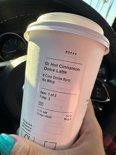 A close-up shot shows a white Starbucks coffee cup held in a person's hand, resting near a car's steering wheel. The cup's label indicates the drink is a "Gr Hot Cinnamon Dolce Latte" with "6 Cinn Dolce Syrp" and "No Whip." The receipt portion of the label states "Item: 1 of 3", "Total: 3", and "7:11 AM 15-Apr-2026." The cup also has a recycling logo and text stating "This cup has be redesigned wi 30% recycled." The hand holding the cup has a nail with a light blue polish. The steering wheel and dashboard of the car are visible in the background. This is a drive thru drink. It is unknown if the city is in Huntington Beach.