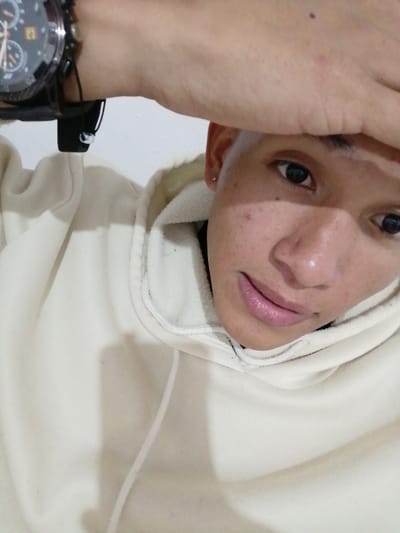 A person wearing a light beige hoodie poses for a close-up selfie. Their right hand is raised, covering part of their forehead, adorned with a large, black sports watch. The individual has dark eyes, short hair, and a small earring. The background appears to be a plain, light-colored wall. The image is well-lit, highlighting the person's features.