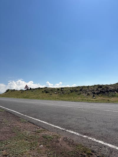 The image displays a roadside scene with a clear blue sky and a gently sloping, grassy embankment on the left. The embankment is dotted with rocks and sparsely covered with vegetation. A paved road runs along the right side of the frame, with a white line marking the edge. The road surface appears somewhat worn, with visible cracks.

A triangular road sign with a red border and a black symbol depicting a sharp curve to the right is positioned on the embankment, close to the roadside. Fluffy white clouds are scattered across the sky, suggesting fair weather. The sunlight indicates it is daytime. The overall setting appears to be a rural or semi-rural area, possibly in Armenia, given the location context provided. No people are visible in the image. The focus is on the natural landscape and the road.