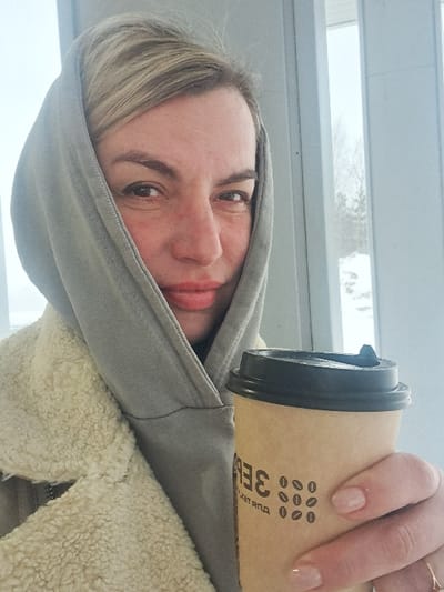 A woman with fair skin and blonde hair, wearing a gray hooded jacket, holds a brown paper coffee cup with a black lid. The cup has a coffee bean design. She is looking directly at the camera with a slight smile. A window with horizontal blinds is visible in the background. She is wearing a ring on her right ring finger. Her nails are painted a neutral shade. She has light-colored jacket underneath the hoodie.