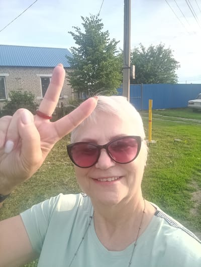 A woman with short, white hair and pink-tinted sunglasses smiles as she holds up two fingers in a peace sign. She is wearing a light blue t-shirt and a dark red ring on her left ring finger. The background includes a brick building with a blue metal roof, green trees, a blue fence, and a yellow and red striped pole. The image appears to have been taken outdoors in Brodokalmak, Russia, given the presence of typical rural Russian elements. The overall lighting suggests it is daytime.