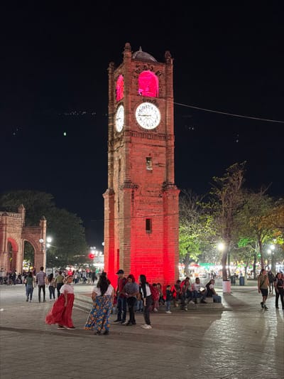An illuminated brick clock tower dominates the night scene, casting a red glow on people seated and standing nearby. The clock face shows 12:00. Above, trees are illuminated, while the background is mostly dark. Below, arches frame a distant view of more people and lights. Two women in colorful skirts stand out in the foreground. The image is vertically oriented.