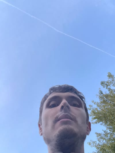 A low-angle, close-up, selfie-style shot features the face of a person against a bright blue sky with a faint contrail. The person's head is tilted upwards, and their eyes are looking directly at the camera. Their hair appears dark and slightly disheveled. There is a light mustache and stubble on their chin. To the right of the frame, the green foliage of a tree is visible against the sky. The overall lighting suggests an outdoor setting during the daytime, likely in Genk, Belgium.