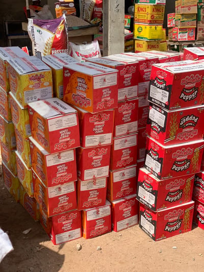 A roadside display of food products shows stacked boxes of "minimie Noodles" in various flavors. The top layer features "Pepper" flavor in red packaging. The middle layers are "Party Jollof" flavor, predominantly red and orange. The bottom layers are "Slurp Chicken" flavor in yellow packaging. In the background, shelves display other food items, including rice and vegetable oil. The overall impression is a small market or roadside shop selling basic food supplies.