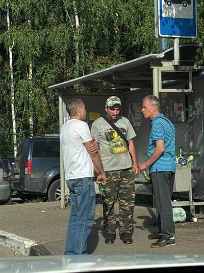 Three men are standing at a bus stop on a sunny day, with trees and a parked SUV in the background. The bus stop has a metal roof and the men appear to be engaged in conversation. One man on the left is wearing a white t-shirt and jeans, holding a green bottle. The middle man is wearing a grey t-shirt with "THAILAND Jungle Tour" printed on it, a camouflage baseball cap, and camouflage pants, also holding a green bottle. The man on the right is wearing a blue and white striped shirt and dark pants, holding a green bottle. Several other green bottles are scattered on the ground near the bus stop. The location is identified as Горбольницы №45, Russia.