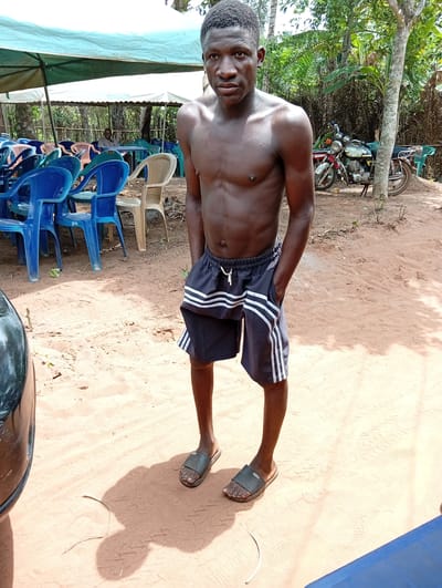 A young Black man stands shirtless on reddish-brown sandy ground, looking directly at the camera with a neutral expression. He wears dark athletic shorts with white stripes on the sides and dark slide sandals, with his hands in his pockets. His skin appears slightly shiny, suggesting warmth or recent activity.
The setting is an outdoor gathering area in Umundugba, Nigeria, characterized by bright sunlight casting strong shadows, indicating midday. In the background, several light blue and white canopies provide shade over numerous plastic chairs in blue, light green, and beige, arranged in rows. A person is visible seated at a table under one of the distant canopies, partially obscured by the chairs.
To the right, against a backdrop of lush tropical trees and foliage, two motorcycles are parked, one red and white, the other green and white. Part of a dark, possibly metallic, object or vehicle is visible in the lower left foreground. The scene conveys a casual, outdoor social or event space in a warm, tropical climate. No discernible text is visible.