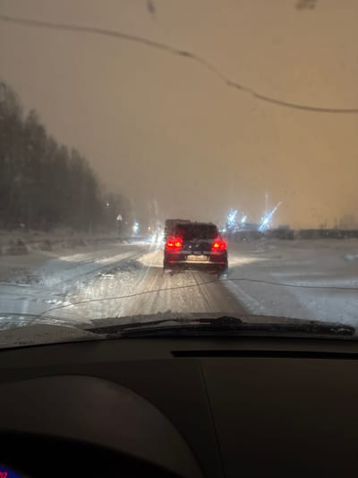 A car is seen through the windshield of another vehicle on a snowy or wet road. The rear car's taillights are illuminated, and other car headlights are visible in the distance. The windshield wipers have cleared a portion of the glass. The overall visibility is limited due to the weather conditions.