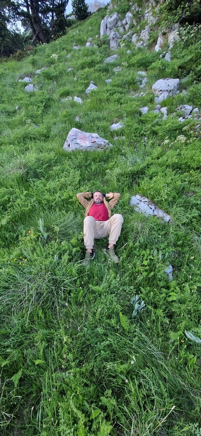 A man is lying on his back in a grassy, sloped area. He is wearing a red t-shirt, a checkered shirt over it, tan pants, and green sneakers. His hands are behind his head, and his eyes are closed. The environment appears to be a natural landscape with grass, rocks, and some trees visible in the background. The location is Ubla, Bosnia and Herzegovina.