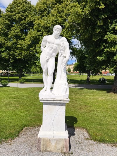In Ekerö kommun, Sweden, a white marble statue of a muscular, nude male figure stands on a cracked marble pedestal. The statue is positioned on a gravel path in front of a lush green lawn, with large, mature trees providing a dense canopy overhead. In the background, beyond the trees, several yellow buildings are visible, and a few people are seated at tables under umbrellas on the grass. The sky is a bright blue with scattered clouds, suggesting a sunny day. The overall scene depicts a public park or garden setting with a classical sculpture as a focal point.