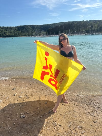 A woman stands on a pebbly beach, holding a bright yellow towel with red lettering. She is wearing sunglasses and a dark bikini top. The towel displays the words "Département" and partially visible "en Vercors". Behind her, the turquoise water of a lake meets the shore, and a line of sailboats is anchored in the distance. A lush, tree-covered hillside forms the backdrop under a clear blue sky. The location is specified as the city of Maisod, France, likely on the Lac de Vouglans. The scene captures a sunny day by the water, with the woman posing for the camera.