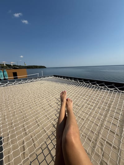 The image displays a first-person perspective, showcasing the bare lower legs and feet of a single individual resting on a large white net hammock. The hammock, stretched over a light tan sandy surface, exhibits a grid pattern with distinct shadows. Beyond the hammock, a calm, dark blue body of water extends to a distant green treeline on the horizon. The sky above is clear blue with sparse white clouds. To the left of the frame, a low-profile brown structure is visible, featuring two light blue paddleboards propped vertically against it and multiple white sun umbrellas. A yellow and blue Ukrainian flag is flying on a pole adjacent to the structure. This scene is located at a recreational facility on the Kyiv Reservoir, in Hlibivka, Kyiv Oblast, Ukraine.