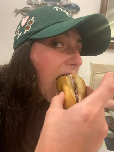 A woman wearing a green baseball cap, adorned with pins, is captured in a close-up shot as she eats a sandwich. The sandwich appears to have a light-colored filling and is held up to her mouth with her hand. The background includes a white wall and an art piece composed of silver fish shapes. Her dark hair frames her face as she looks directly at the camera.