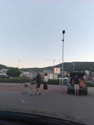 In the city of Prachatice, Czechia, on a paved parking lot, a man in a red cap and athletic shorts pushes a shopping cart with two people visible inside it. Beside him, a woman in a white top and dark skirt walks, her feet bare. To the right, a man in a blue shirt and light shorts unloads an orange box from the open trunk of a dark SUV. The background features a building with a red and white "Kaufland" sign, streetlights, and a wooded hill. The sky is a pale blue, indicating dusk or early morning.
