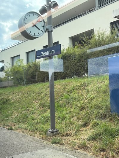 A vertical shot captures a clock and a signpost outside a modern building. The clock, a large, white analog timepiece with black markings and red and black hands, hangs from a metal pole. To the right of the clock, a dark blue rectangular sign is attached to the same pole. The sign reads "Zentrum" in white, bold, sans-serif lettering, with "Meggen" written in smaller white text below it. The background reveals a building with light-colored walls and dark-trimmed windows, set against a backdrop of lush green grass and trees. The scene appears to be in daylight, with diffused lighting suggesting an overcast or partly cloudy sky. The overall impression is one of a public space, possibly a train station or bus stop, in a well-maintained urban or suburban environment. The image is captured from a slightly low angle, looking up towards the clock and sign.