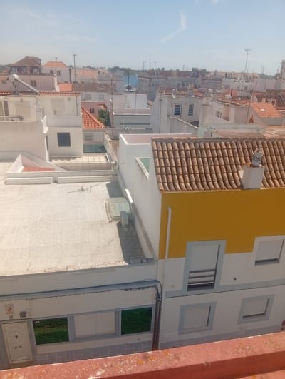 The image captures a view of the rooftops in Vila Real de Santo António, Portugal, under a clear, bright sky. The architecture features a consistent style of white buildings with red-tiled roofs, interspersed with occasional colorful accents, such as a prominent yellow facade on one building in the foreground. The density of buildings suggests an urban or semi-urban setting. The foreground shows a section of an orange, weathered wall or railing. On the buildings, various elements are visible, including windows with white shutters, ventilation units, satellite dishes, and chimneys. The overall scene is one of a sun-drenched day in a European coastal town.