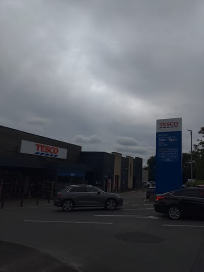 The image shows the exterior of a Tesco supermarket in Magherafelt, United Kingdom. The sky is overcast with grey clouds. In the foreground, a dark-colored car is parked. A grey SUV is driving on the road in front of the supermarket. The Tesco building has a large white sign with red lettering that says "TESCO". A tall blue sign with the Tesco logo and opening hours is also visible. The supermarket appears to be open, as indicated by the signage. The area around the supermarket is paved, with some trees and streetlights visible in the background.