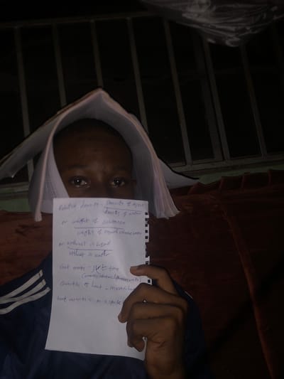 A person is lying on a surface that appears to be a bed or couch, holding a piece of paper with handwritten notes. The notes on the paper relate to physics concepts, including "Relative density", "Heat energy", "Quantity of heat", and "heat capacity." The person is positioned near a window with metal bars. The location is Atani, Nigeria.
