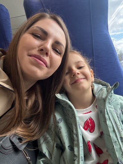 A selfie taken in what appears to be public transportation shows an adult female and a young girl. The adult female is lying on a blue seat, with her head close to the camera. She has long, brown hair and is wearing a beige jacket. The young girl is positioned slightly behind and above the adult female, also lying down. She is wearing a white sweater with red heart drawings and a patterned green jacket. The background includes a glimpse of the outside through a window. A black handbag is partially visible near the adult female. No evidence indicates the location is in Покровка, Russia.