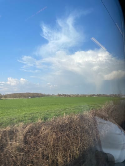 The image depicts a rural landscape within Hochfelden, France, viewed from inside a moving vehicle. The immediate foreground is characterized by motion-blurred, dry, brown vegetation, and reflections on a window pane, including a partial white object at the bottom right. A large, vibrant green field occupies the midground. Along the distant edge of this field, a line of deciduous trees, currently devoid of foliage, suggests late winter or early spring. A white cargo van is parked or driving on an unseen road or track among these trees. The sky above is divided: the left displays a clear light blue with scattered white cumulus clouds and a faint, linear contrail. The right side features a dense accumulation of dark grey cumulonimbus clouds, from which visible shafts of precipitation descend towards the horizon. No individuals are present in the observable landscape.