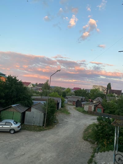 This is an outdoor, eye-level, medium shot photograph taken in the city of Khust, Ukraine. The image depicts a rural or semi-rural street lined with numerous small garages, some with corrugated metal siding, others with wooden doors, and some appearing weathered or in disrepair. A silver sedan is parked in front of a green garage. Vegetation, including trees and bushes, is abundant throughout the scene. In the background, several residential buildings, including what appears to be a multi-story apartment building, are visible amongst the trees. The sky is a soft blue with scattered clouds tinged pink by the setting sun, creating a peaceful sunset atmosphere. A street lamp is present, and utility lines are visible strung across the frame. The ground is a dirt or gravel road. A set of concrete stairs with a metal railing is in the foreground on the right side of the image, with a bicycle wheel visible at the bottom.