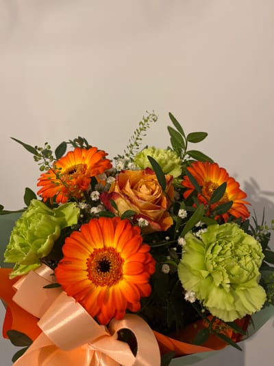 A close-up, static, indoor photograph depicts a floral bouquet. The bouquet features multiple orange gerbera daisies, several light green carnations, a single prominent rose exhibiting orange, peach, and reddish-pink tones, white baby's breath, and various green foliage elements. The arrangement is presented in a wrapping of green and orange paper, secured at the base with a peach-colored satin ribbon tied into a bow. The background is a plain, light beige surface, showing subtle shadowing from the bouquet. No individuals are visible. This scene is located in the city of Iława, Poland.