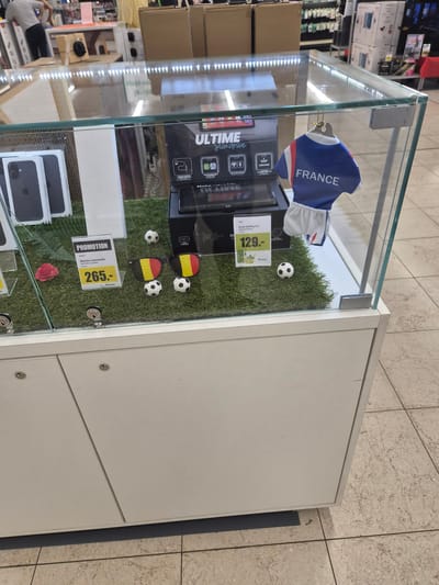 This media file captures a retail display case showcasing electronic devices and accessories. The display is oriented vertically, with the bottom of the image appearing to be on the left side. The scene is set in a store, as indicated by the tiled flooring and the presence of other merchandise in the background.
The display case itself is made of glass and metal, revealing a collection of items arranged on artificial grass. Prominently featured are several boxed Apple iPhones and an Apple Watch. Other electronic devices, possibly tablets or portable media players, are also visible within the case, along with packaging for a "Slimdrive" and an "Ecran Carplay 8.1".
Decorative elements include miniature soccer balls and a red rose, suggesting a thematic promotion, possibly related to sports or a special event. A miniature French national football team jersey, emblazoned with "FRANCE," further reinforces this theme. Price tags are visible, with one indicating "129.-" and another "265.-" for what appears to be a connected watch.
Text within the display includes brand names like "APPLE," "SWATCH," and "Slimdrive," as well as promotional phrases like "PROMOTION" and product details. The overall impression is that of a brightly lit retail environment aimed at attracting customers with deals on consumer electronics. The specific location context of Käerjeng, Luxembourg, suggests this is a store within that region. No people are clearly identifiable in the foreground of the display, though a person can be seen in the background, to the left, interacting with merchandise.
