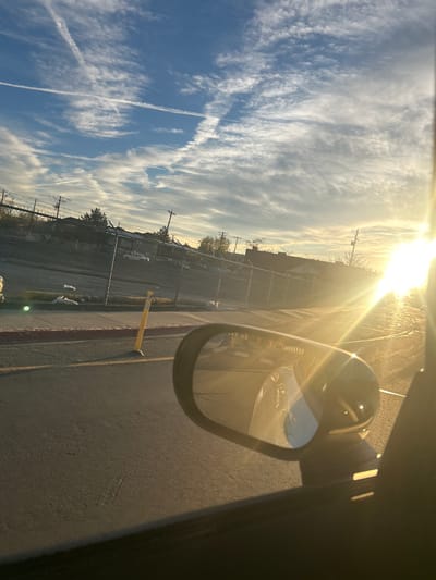 The image depicts a street scene in an urban area of Reno, Nevada, United States, viewed from the passenger side of a vehicle during daylight with the sun low on the horizon, causing significant lens flare on the right.

The sky is predominantly blue, covered with scattered white cirrus and cirrocumulus clouds. Two distinct airplane contrails form an 'X' pattern across the upper left sky. The sun is positioned to the far right, emitting a strong golden light that flares across the right half of the image.

In the foreground, the dark interior trim of a vehicle's window frame is visible along the left and bottom edges. A passenger side mirror is prominent in the lower right, reflecting a portion of the vehicle's white exterior and the road surface behind. The asphalt road surface occupies the lower half of the view.

Immediately adjacent to the road, a red-painted curb borders a concrete sidewalk. A single yellow, cylindrical bollard with a white conical base stands slightly angled on the sidewalk to the left of the mirror. Behind the sidewalk, a dark chain-link fence, approximately six to eight feet high, topped with two strands of barbed wire, stretches horizontally across the midground. Beyond the fence, a dirt or grassy embankment is visible, containing scattered white plastic bags or debris. A circular green lens flare appears on the ground near the fence line.

In the background, beyond the fence and embankment, several vehicles are parked, including a white pickup truck. Further back, multiple commercial or residential buildings are present. One building features a dark sign with light-colored text clearly reading "DAY CARE." Utility poles and numerous power lines are visible throughout the background, with sparse trees and bushes interspersed among the structures, some appearing dormant. No people are visible.