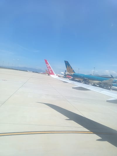 The image shows a tarmac at Cam Ranh International Airport (CXR) in Bắc Cam Ranh, Vietnam. Several airplanes are parked on the tarmac under a clear blue sky. In the foreground, the wing of an airplane casts a shadow on the ground. One airplane in the mid-ground is predominantly blue with a yellow emblem on its tail and the text "VN-A896" visible. Another airplane to its left has a red tail with white text that reads "Air Asia". In the background, distant mountains are visible, along with airport lights and other aircraft. The tarmac is paved with concrete and marked with yellow and white lines.
