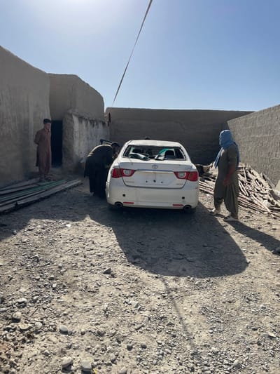 This image depicts an outdoor scene in Spin Boldak, Afghanistan, characterized by rudimentary construction and a damaged vehicle under clear skies. A white Toyota Mark X sedan is centrally positioned, facing away from the viewer. Its rear window is completely shattered, with visible glass fragments inside the vehicle. The license plate area is vacant.
Three males are present. On the far left, a male in a light brown shalwar kameez stands idly near a dark doorway of an adobe-style structure. Directly behind the white sedan, another male, dressed in a dark shalwar kameez, is bent over, appearing to inspect or work on the vehicle's rear. To the right of the vehicle, a third male, wearing a light brown shalwar kameez and a light blue head covering, stands observing the car.
The environment consists of unpaved, gravelly ground. Low-rise adobe and mud-brick walls and structures form the background. To the right of the car, a pile of irregular wooden debris or building materials is accumulated against a wall. A thin, light-colored strap or rope extends diagonally across the upper portion of the frame. Shadows are distinct and sharp, cast by the individuals and the vehicle, indicating direct overhead sunlight.