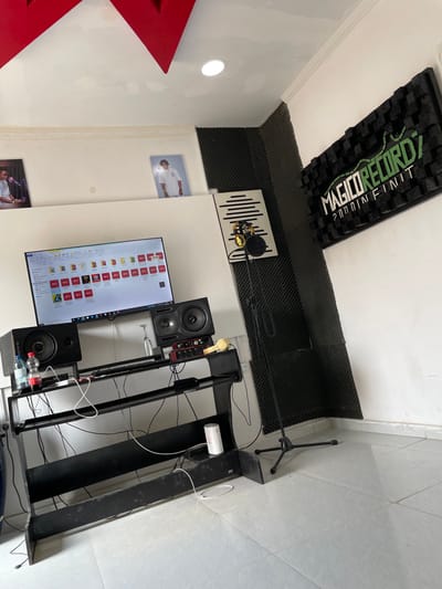 This image presents an indoor recording studio or music production setup. The room features white walls, with extensive black acoustic foam panels covering portions of the walls and along the wall-ceiling boundaries for sound treatment. A prominent, large rectangular acoustic panel with raised patterns and green text, "MAGO RECORDZ 200 INFINITE," is affixed to the ceiling.
At the center of the workspace, a professional microphone with a pop filter is mounted on an arm, positioned in front of a white acoustic panel featuring a vertical wave-like design. Below it, a black desk or workstation holds a large computer monitor displaying numerous icons, some labeled "SOLD," alongside two studio monitor speakers, one branded "M-AUDIO." A red "Focusrite" audio interface and other audio equipment are also arranged on the desk. Numerous cables are visible, connecting the various electronic components. On a lower shelf of the desk, two water bottles and a white cylindrical object are present.
To the right, a black microphone stand with a tripod base rests on the light-colored tiled floor. The white walls also feature decorative elements, including two framed pictures; the upper one shows a person in a white sports jersey. A bright circular recessed light is visible on the left wall, alongside a large red architectural element partially seen on the far left. The setting is equipped for audio recording and production, though no activity is currently in progress, and no individuals are visible. The lighting appears to be artificial.