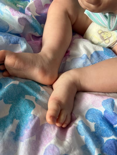 The image displays a close-up view of a baby's lower body, specifically the legs and feet, resting on a colorful patterned blanket. The scene is set in Dubovka, Russia. The baby's skin is smooth and light-toned. A disposable diaper, light yellow or cream in color, is visible around the waist, adorned with a repeated cartoon monkey face pattern. Part of a light blue and white striped fabric, possibly a swaddle or clothing, is also visible near the diaper.

The baby's legs are bent, and the feet are relaxed, resting on the fabric. The sole of one foot is partially visible, showing typical creases. The fabric beneath the baby features a vibrant, abstract floral or blotchy pattern in shades of blue, purple, and pink/lavender. The lighting suggests it is daytime, providing soft illumination. The baby appears to be resting peacefully. No discernible text is visible on the diaper or any other object in the frame.