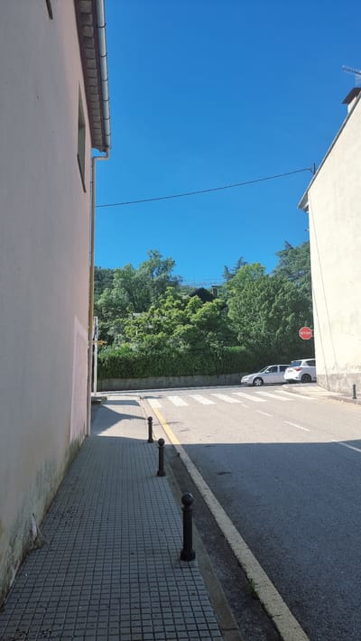 This is a medium-angle, full shot of a street scene in Castellfollit de la Roca, Spain. The image is dominated by a clear blue sky and a bright sunny day. In the foreground, on the left, is the side of a cream-colored building with a metal gutter system. A gray tiled sidewalk with several black bollards runs alongside the building. To the right of the sidewalk is a street with a pedestrian crossing. A yellow line marks the edge of the sidewalk. Further down the street, two cars are parked, a silver sedan and a white SUV. On the right side of the street, another cream-colored building is partially visible. Beyond the cars and buildings, a dense green forest covers a hillside, with some houses or structures visible through the trees. A red octagonal stop sign is mounted on a pole at the edge of the road. The lighting suggests it is midday, with strong shadows cast by the buildings and bollards.