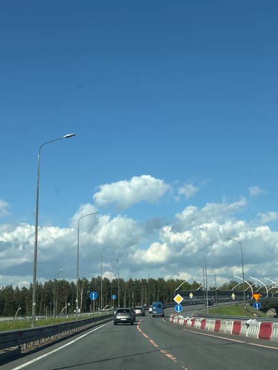 The image depicts a view from a vehicle moving on a road in Beloostrov, Russia. The right side of the frame shows a highway with guardrails, road markings, and signage. Traffic includes a blue truck and other vehicles. On the other side of the highway, there is a line of trees. Streetlights run parallel to the highway. The sky occupies the left portion of the image, featuring a bright blue expanse juxtaposed with white clouds.
