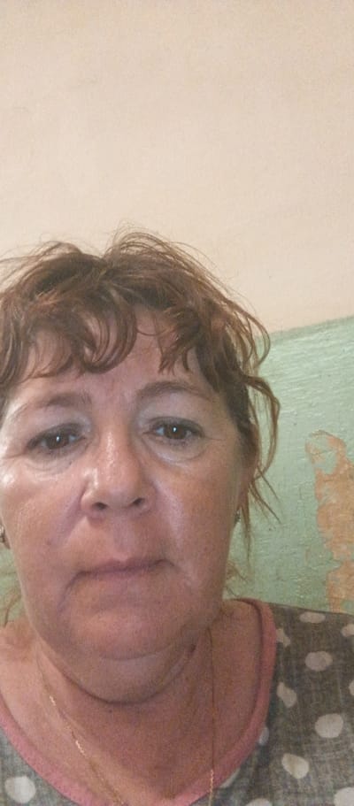 A close-up, front-facing selfie shows a middle-aged woman with brown, slightly messy, shoulder-length hair. She has brown eyes, a fair complexion with some visible skin texture, and is wearing a gold necklace. Her top has a pink neckline and a pattern of white polka dots on a grey background. The background consists of a plain, light-colored wall in the upper portion and a textured, green-painted wall with peeling paint in the lower right. The image appears to have been taken indoors. There is no information in the image to indicate the location in Beregovoe, Ukraine.