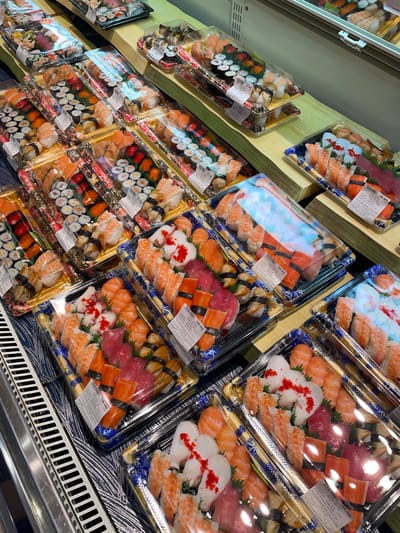 The image displays a retail setting, likely a grocery store or market, with a prominent display of pre-packaged sushi. The sushi platters are arranged in clear plastic containers and are stacked on wooden shelves and in a refrigerated display case. The products vary in size and assortment, featuring different types of sushi rolls and nigiri, with visible ingredients like salmon, tuna, shrimp, and imitation crab. Some packages have labels with text and pricing information. The environment appears to be indoors, with overhead lighting and a clean, organized presentation of merchandise. There are no people visible in the frame. The bottom left corner of the image shows a section of a metal refrigerated display case with horizontal grating. The overall impression is a well-stocked food retail display focused on sushi. The mention of Nghĩa Trụ Commune, Vietnam, suggests the location of this retail establishment.