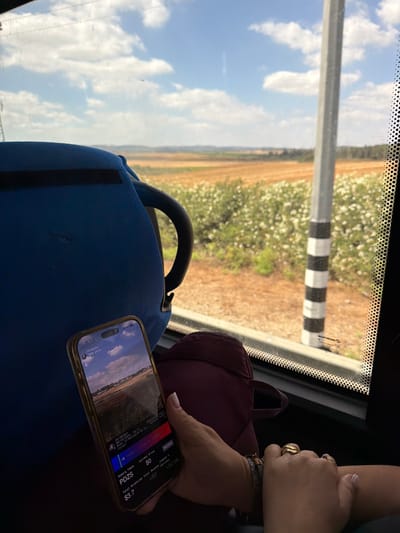 A person's hands are visible in the foreground, resting on their lap or armrest. They are wearing several rings on their fingers. In the center of the image, a smartphone is held, displaying a screen with what appears to be some form of digital interface or application. The phone is displaying the time as "10:14" and a partial view of a landscape with clouds. The background shows a view from inside a vehicle, likely a bus, looking out a window. The window looks out onto a rural landscape with fields of crops or vegetation, a blue sky with clouds, and a striped pole outside. Based on the phone's screen information, the location is the city of Modi'in-Maccabim-Re'ut, Israel. A blue bag is visible on the left side of the image, and a portion of a burgundy bag is also present.