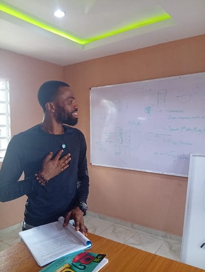 The scene depicts an adult male inside a room in Evbogida, Nigeria. The man, exhibiting dark skin, short dark hair, and a beard, is positioned in profile, facing right, with his mouth open in an apparent smile. He wears a black long-sleeved shirt, a dark necklace with a blue and white circular pendant, a brown beaded bracelet on his left wrist, and a dark watch on his right wrist. His left hand is placed flat on his chest, while his right hand rests on an open white binder with a blue spine, situated on a light brown wooden table in the foreground. Below the binder, a colorful book or magazine cover displaying a red rotary phone and "80%" text is partially visible.
The room features peach-colored walls and a light-colored tiled floor. A large white whiteboard is mounted on the back wall, covered with blue marker diagrams and text, including "DB," "Voltmeter," "Fuse, Capacitor, Semi," "Generator, Meters, Relay," and "Plier, Screwdriver." A smaller, vertically oriented white board or framed item is partially visible on the floor to the right of the large whiteboard. On the left wall, a white-framed gridded window is present. The white ceiling incorporates a recessed rectangular section with green LED strip lighting and a single circular recessed light fixture.