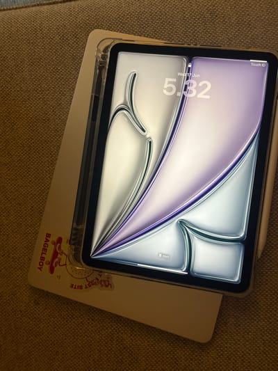 The image displays a close-up, top-down view of an iPad and a laptop on a textured, brown surface, likely a couch or chair. The iPad is in a clear protective case and rests on top of a white laptop. The iPad's screen is illuminated, showing a lock screen with the time "5:32" and the date "Wed 17 Jun". The wallpaper on the iPad features an abstract, iridescent design with purple and silver hues, creating a sense of depth and shine. A stylus, possibly an Apple Pencil, is tucked into the top edge of the iPad case.
The laptop has a sticker on its surface with a cartoonish design of a bagel character riding a skateboard. The text on the sticker reads "BAGELBOY" and "BEST BITE" in a playful font. There are also smaller inscriptions like "EST 2023" and "AMS NL".
The lighting suggests it might be daytime, but the ambient light is soft, casting subtle shadows. The overall scene conveys a sense of everyday technology use, perhaps in a home or casual setting. There are no people visible in the image. The context of Aarhus, Denmark, does not directly influence the visual elements of the image itself, but provides a geographical location for the scene.