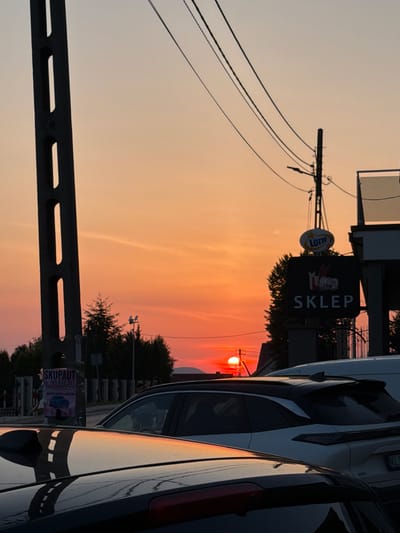 This is a photograph taken at dusk in the city of Mordarka, Poland, featuring a vibrant sunset. The sky transitions from a pale orange at the top to a fiery red and pink near the horizon, where the sun is setting. In the foreground, the reflections of streetlights and the sky are visible on the polished surfaces of cars, suggesting a street scene.

Prominently displayed is a black sign with "SKLEP" in white capital letters, indicating a shop, and above it, a "LOTTO" logo, suggesting a lottery retailer. A tall, dark utility pole with cutouts stands to the left of the frame, with power lines stretching across the sky. Another utility pole with a street lamp is visible further to the right, near the shop sign.

To the left of the utility pole, a poster is attached, featuring text in Polish, including "SKUP-AUT" and a phone number. In the middle ground, trees form a dark silhouette against the colorful sky. Several cars are parked or stopped, with their shapes visible against the backdrop. The overall atmosphere is one of a typical evening in a Polish town, with the dramatic sunset adding a striking visual element.
