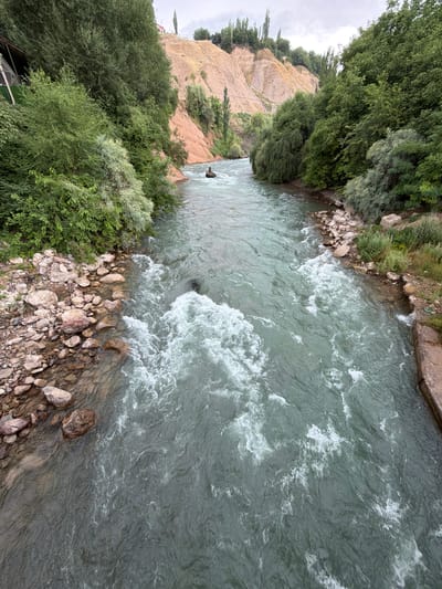The image shows a wide, fast-flowing river with turbulent water. The river is surrounded by lush green trees on both banks, with a steep, sandy hillside in the background. In the middle of the river, a person is visible in a dark object, appearing to be navigating the rapids. On the left bank, there is a small, covered structure with a green roof. The riverbed appears to be rocky, with stones visible along the edges. The sky is overcast, suggesting a cloudy day. The scene depicts a natural environment with a recreational activity taking place. The location is identified as Khumsan, Uzbekistan.