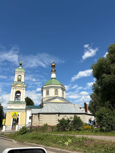 The image shows a bright, sunny day in Petrovskoye, Russia, with a clear blue sky filled with fluffy white clouds. The scene is dominated by two onion-domed church towers, characteristic of Russian Orthodox architecture. The towers are painted in vibrant yellow with green roofs and white trim. One tower features a clock face showing approximately 11:30. The foreground is partially obscured by a car, suggesting the photo was taken from a vehicle.

To the right of the church buildings, a narrow paved road runs alongside a patch of green grass and a dark metal fence. Beyond the fence, a yellow brick building stands, with some trees and vegetation growing around it. In the distance, more trees can be seen, some with dense green foliage.

On the roadside, there are a few yellow traffic signs with red borders. One sign depicts figures of people in a triangle, possibly indicating a pedestrian crossing or a warning about children. Another sign, partially visible, also has a yellow background and a red border with a symbol inside. A few people are visible in the background near the yellow brick building, some appearing to be interacting with objects or cars.

The overall impression is of a pleasant, typical day in a Russian town, with the striking church architecture being the central focus. The sunlight suggests it is late morning or early afternoon.